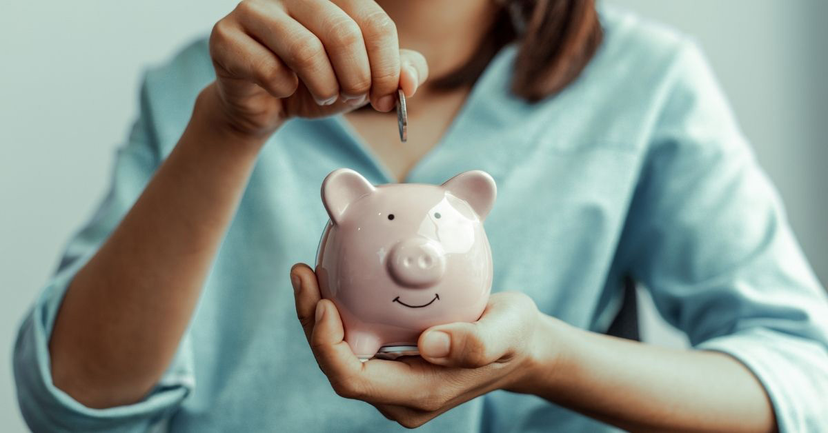 Image of a woman holding a smiling piggy bank, and she is adding a coin